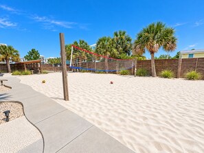 A sandy beach volleyball court with a wooden post, net, and palm trees in the background. - A Beach Life 3A | 3BR Beach Getaway + Pool (Gulf Shores)