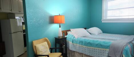 Un dormitorio con paredes verde azulado, una cama con ropa de cama estampada, una silla con cojín, una mesita de noche con lámpara y una ventana con persianas.