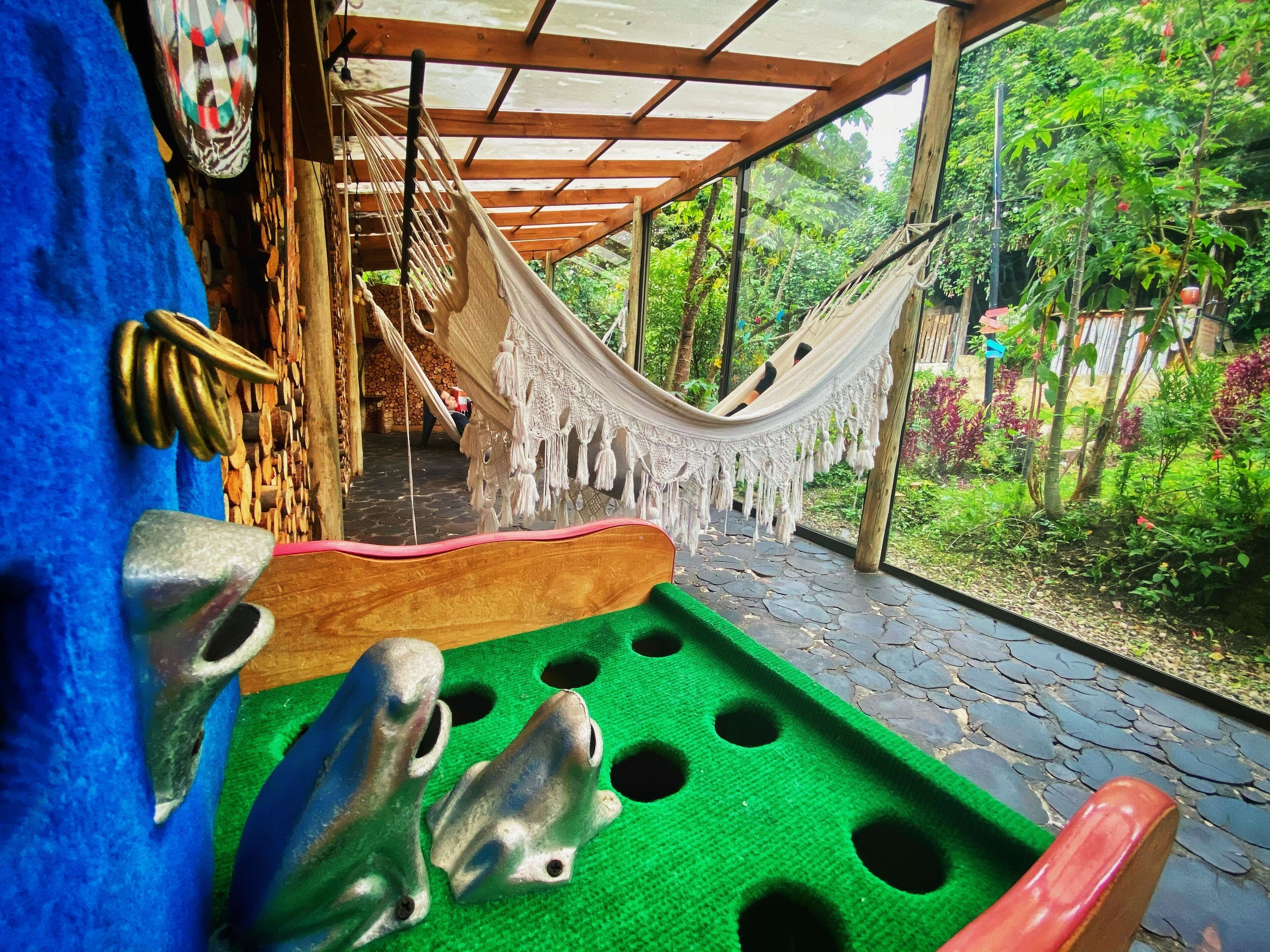 Una hamaca bajo una pérgola de madera, un tapete de juego verde con agujeros, y un interior rústico con suelo de piedra.