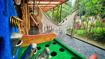 Una hamaca bajo una pérgola de madera, un tapete de juego verde con agujeros, y un interior rústico con suelo de piedra.