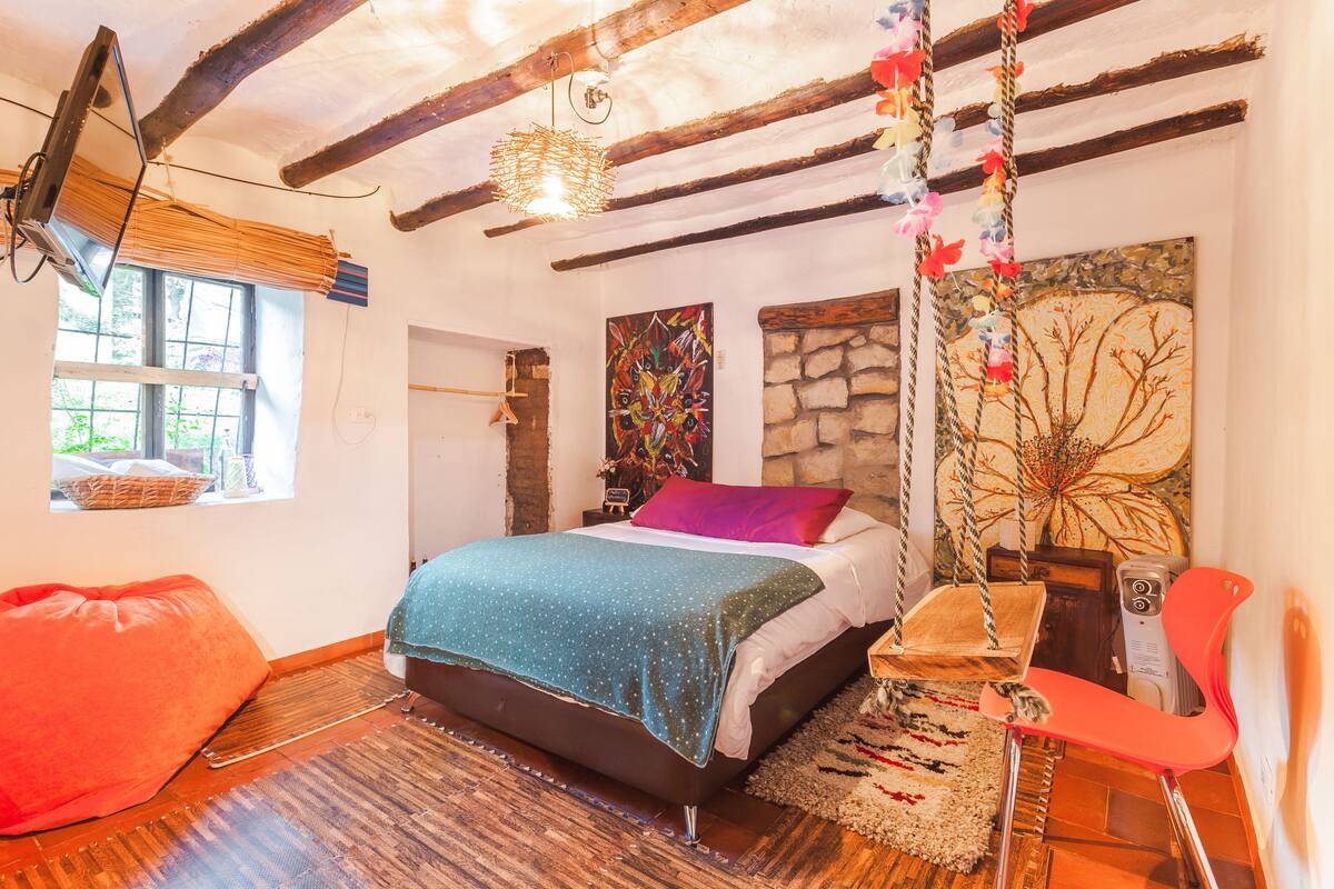 A bedroom with a bed, a red chair, a hanging swing, and a stone wall.