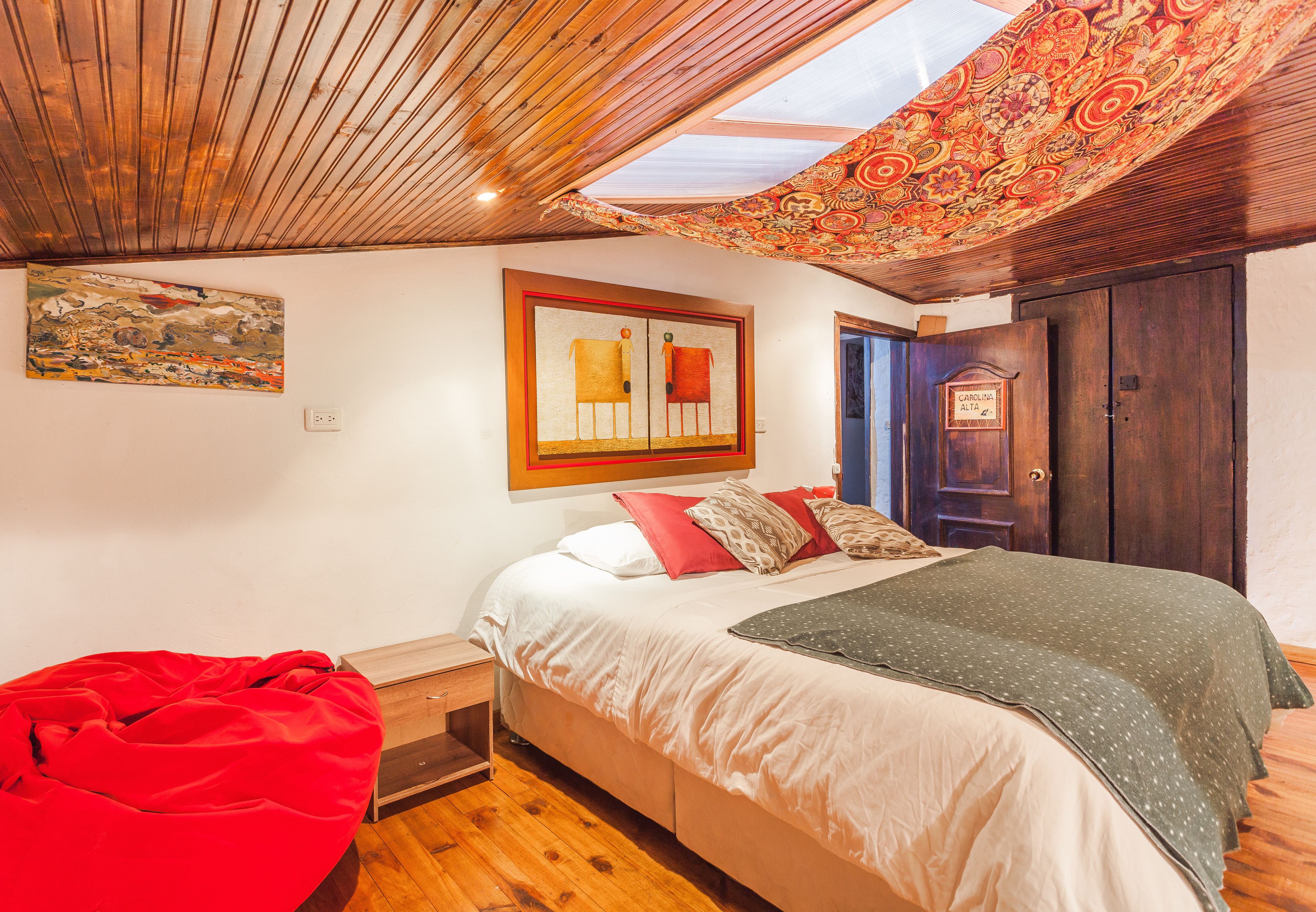 Une chambre avec un lit, un pouf rouge en forme de haricot, un plafond en bois et une porte portant l’inscription « CAROLINA ALTA ».
