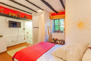 A bedroom with a bed, a TV mounted on the wall, a window with a view of greenery, and a red wall with a sun drawing. - Casa Agrreste Bed & Breakfast (Sopó)