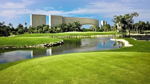 A golf course with a water hazard, palm trees, and a large building with a unique arch structure in the background. - TAO Modern Retreat (Akumal)