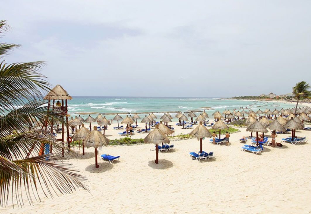 Una playa con numerosas sombrillas de paja y sillas de descanso azules.