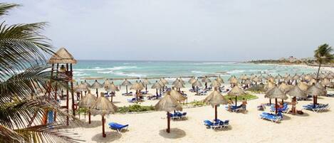 Una playa con numerosas sombrillas de paja y sillas de descanso azules.