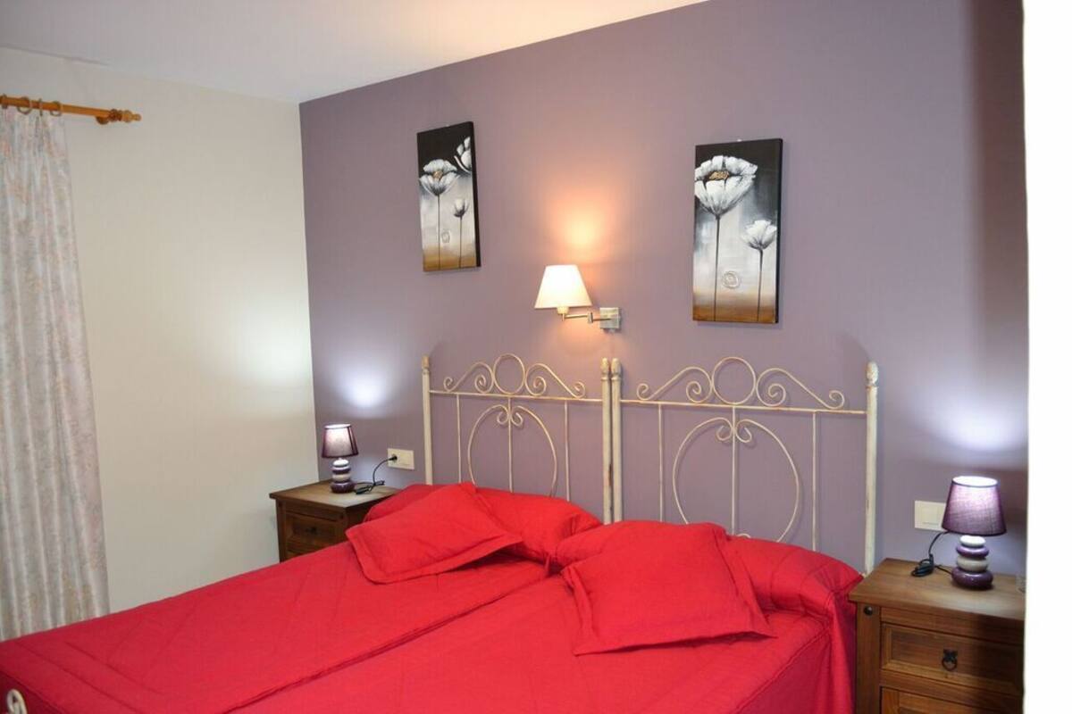 Une chambre à coucher avec un lit en métal, une literie rouge, deux lampes fixées au mur et deux tableaux représentant des motifs floraux.