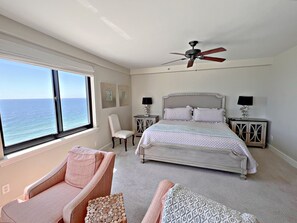 Une chambre avec un grand lit, un ventilateur de plafond, une chaise, une table de chevet et une vue sur l’océan.