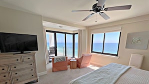 Een hotelkamer met een bed, een televisie, een plafondventilator, een balkon met een stoel en uitzicht op de oceaan.