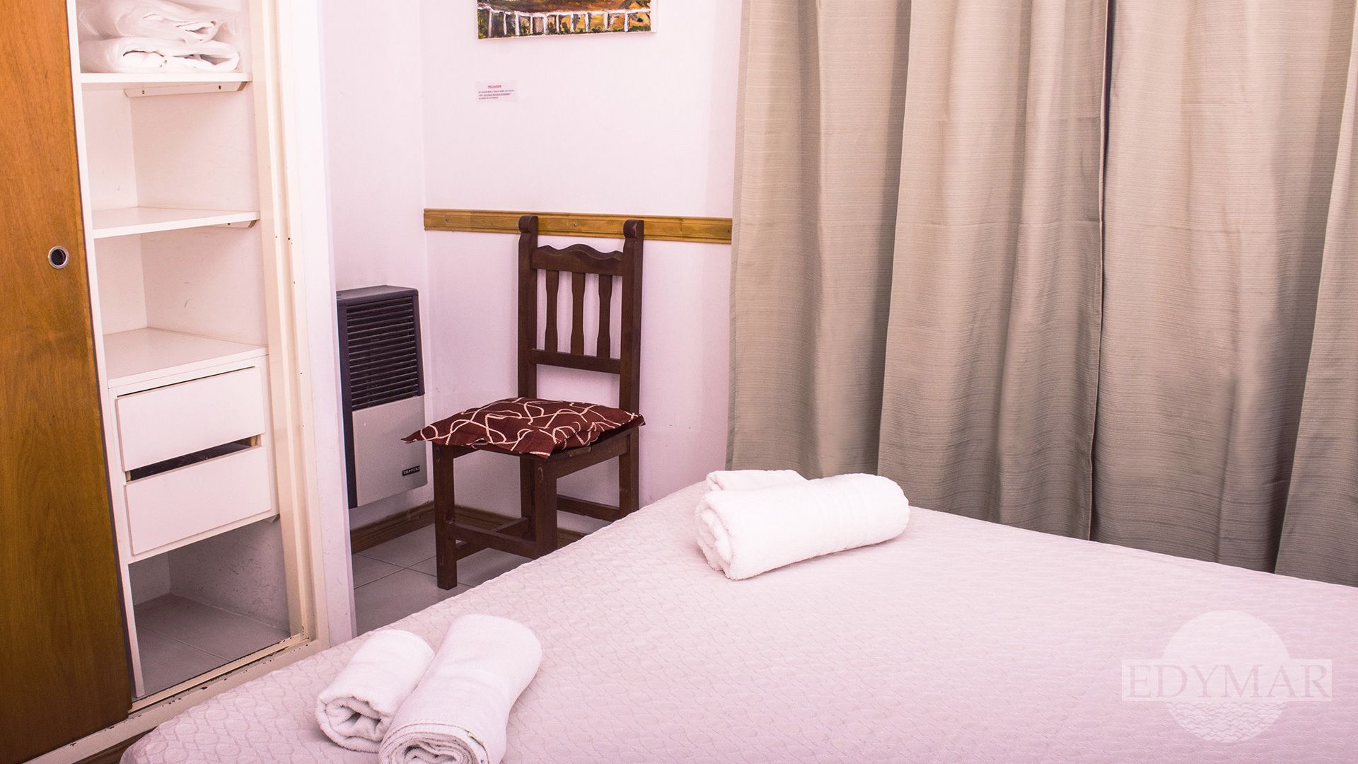 Quarto de hotel com cama, uma cadeira com almofada estampada, guarda-roupa e uma obra de arte pendurada na parede.