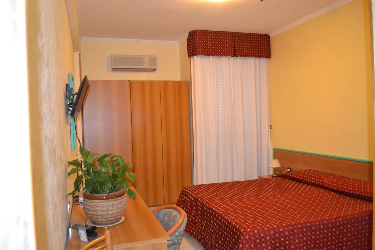 A hotel room with a wooden wardrobe, a bed with a red patterned bedspread, a potted plant, and a desk with a lamp.