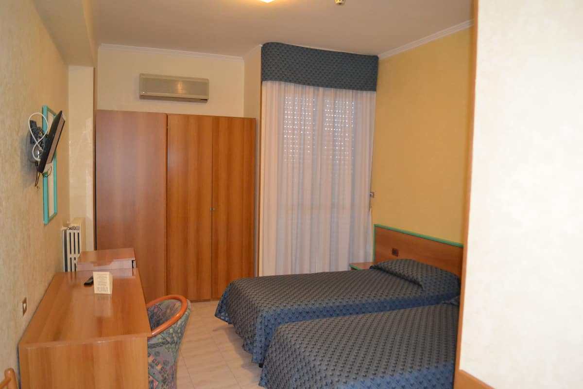 A hotel room with two beds, a wooden desk, a chair, a radiator, and a window with curtains.