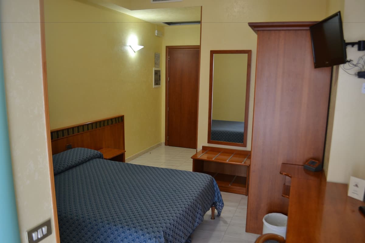 A hotel room with a bed, a wooden bench, a mirror, a television, and a door.