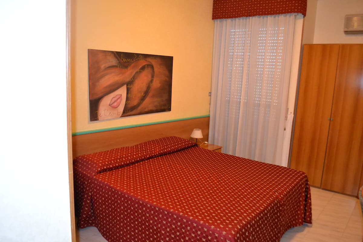 A bedroom with a bed, a nightstand, a lamp, a painting on the wall, and a window with curtains.