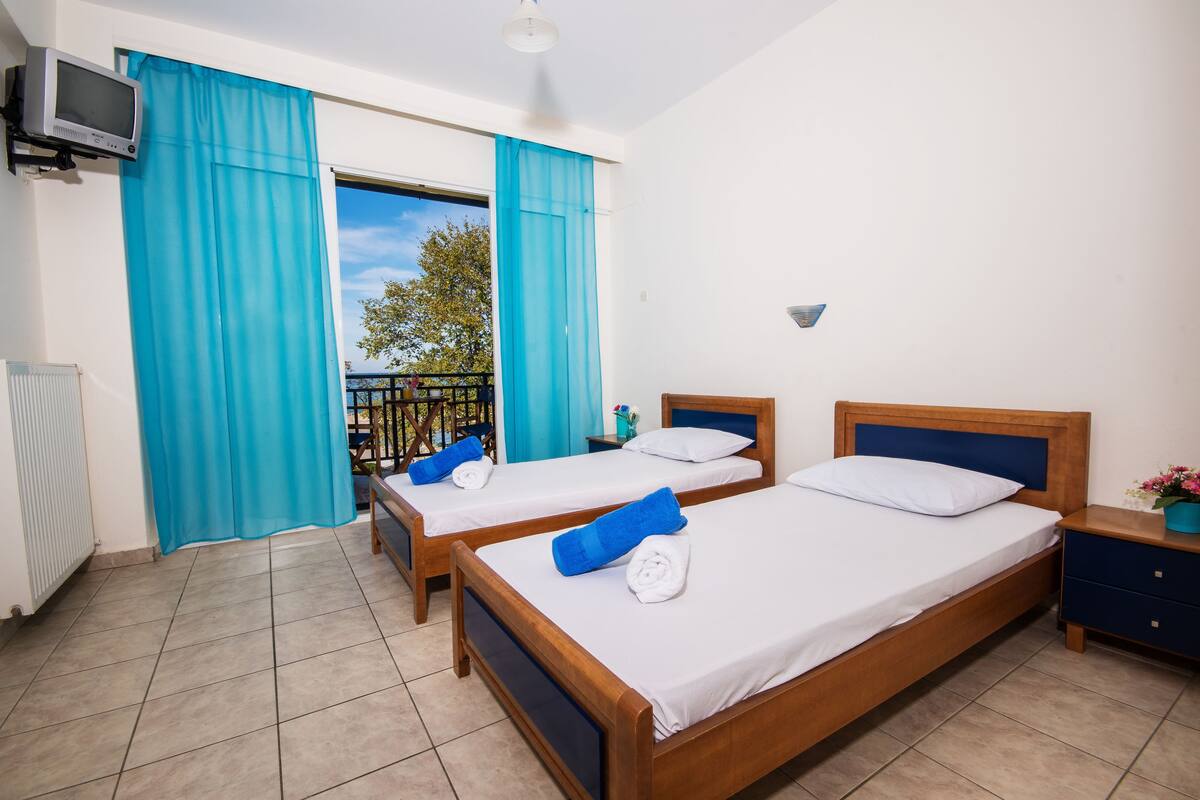 A room with two wooden beds, a TV mounted on the wall, and a balcony with blue curtains.