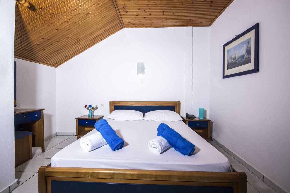 A neatly made bed with two rolled towels, a wooden headboard, a bedside table with a vase of flowers, and a framed picture on the wall.