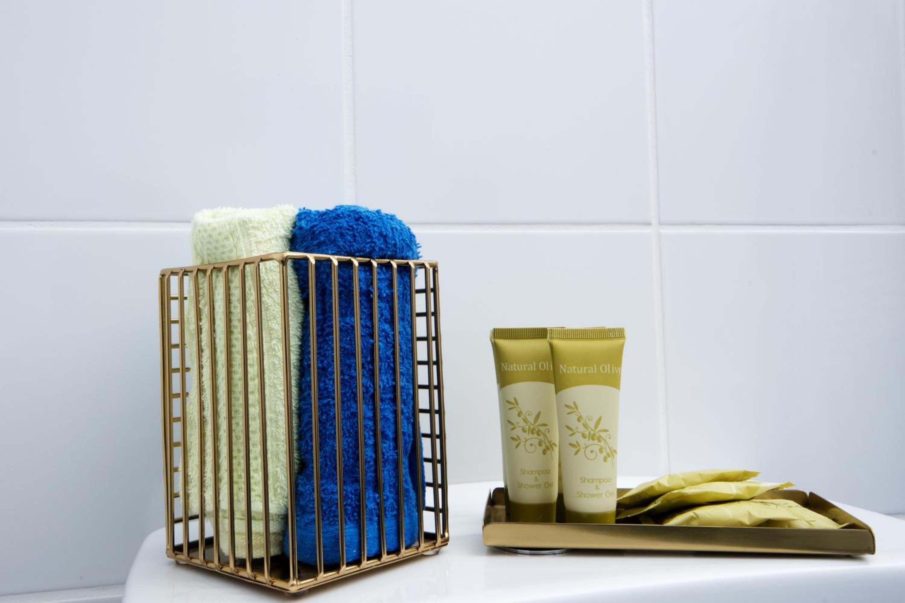 Kylpyhuoneessa on valkoisilla laatoilla päällystetty seinä, kultaisella kehyksellä varustettu kori, jossa on valkoinen ja sininen pyyhe, sekä kaksi luonnollista öljyä sisältävää shampootuubia tarjottimella.