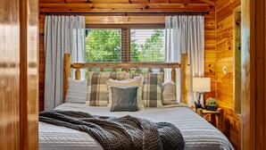 A wooden cabin bedroom with a bed, pillows, a blanket, a nightstand, and a window with curtains. - Luxurious, Romantic, Cozy Cabin/Great Reviews/Close to Town/Clean/Hot tub! (Gatlinburg)