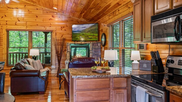 A cozy cabin interior with a stone fireplace, wooden walls, a leather sofa, a kitchen with modern appliances, and a large window offering a scenic view. - Luxurious, Romantic, Cozy Cabin/Great Reviews/Close to Town/Clean/Hot tub! (Gatlinburg)