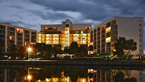 Um hotel com vários andares e varandas, uma piscina refletora à frente e um céu sereno ao entardecer.