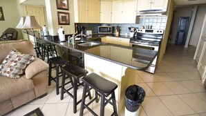 Cozinha compacta com ilha central, banquetas, um sofá com almofada estampada e uma lixeira.