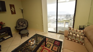 Sala de estar com sofá, mesa de centro, poltrona, televisão e vista para a praia através de uma porta de vidro deslizante.