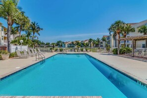 A rectangular swimming pool with a clear blue water, surrounded by lounge chairs and palm trees, with a row of buildings in the background.