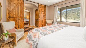 A bedroom with a large bed, a chair, a small table with a plant, wooden barn doors, and a view of the desert.