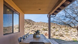 A covered dining area with a long table and chairs, a potted plant, and a view of a desert landscape.