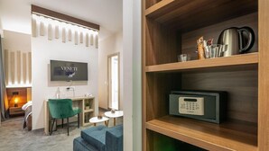 A modern hotel room with a flat-screen TV, a desk with a chair, a sofa, a wooden shelf with a kettle and a coffee maker, and a thermostat. - Veneti Suites (Belgrade)
