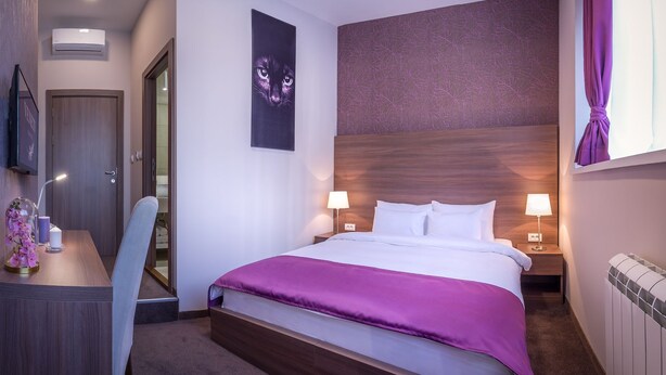 A hotel room with a large bed, two bedside lamps, a headboard, a desk with a chair, a small table with a vase and candles, and a wall-mounted air conditioner. - Veneti Suites (Belgrade)