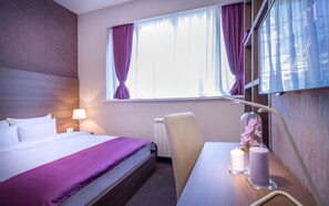 Una habitación de hotel con una cama, un escritorio con lámpara y una ventana con cortinas moradas.