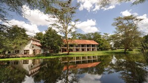 A building with a red roof and white walls, surrounded by trees and a pond reflecting the structure. - Hunters Lodge - Kiboko (Kiboko)