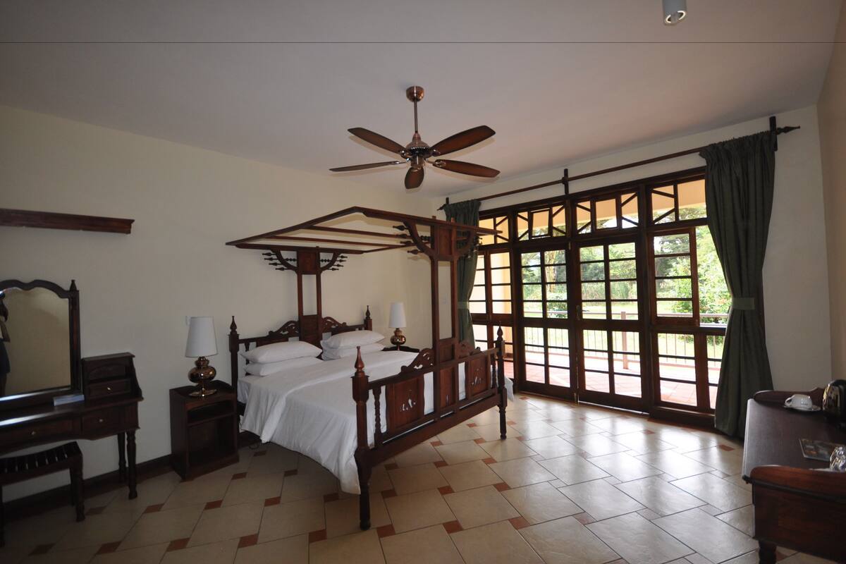 A traditional four-poster bed with a ceiling fan, a large window with curtains, and a wooden nightstand with a lamp.
