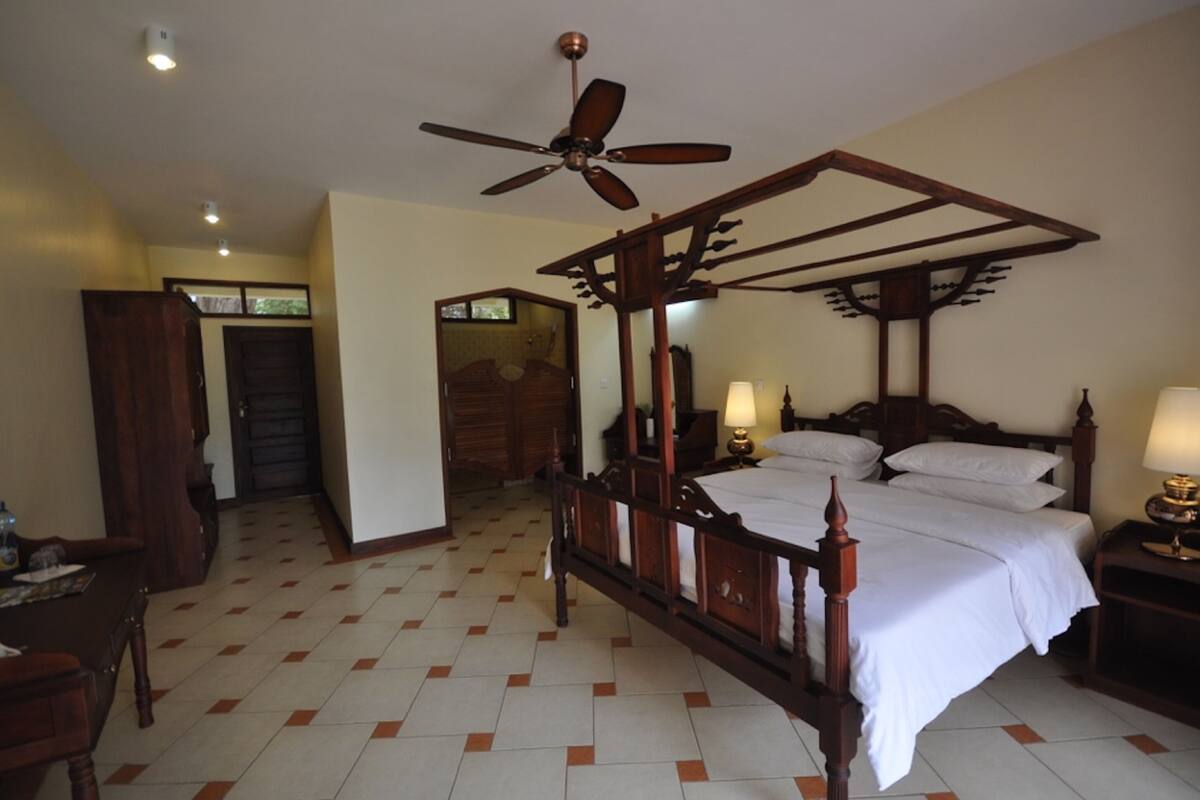 A four-poster bed with a wooden frame and white bedding, a ceiling fan, and a tiled floor with a checkered pattern.