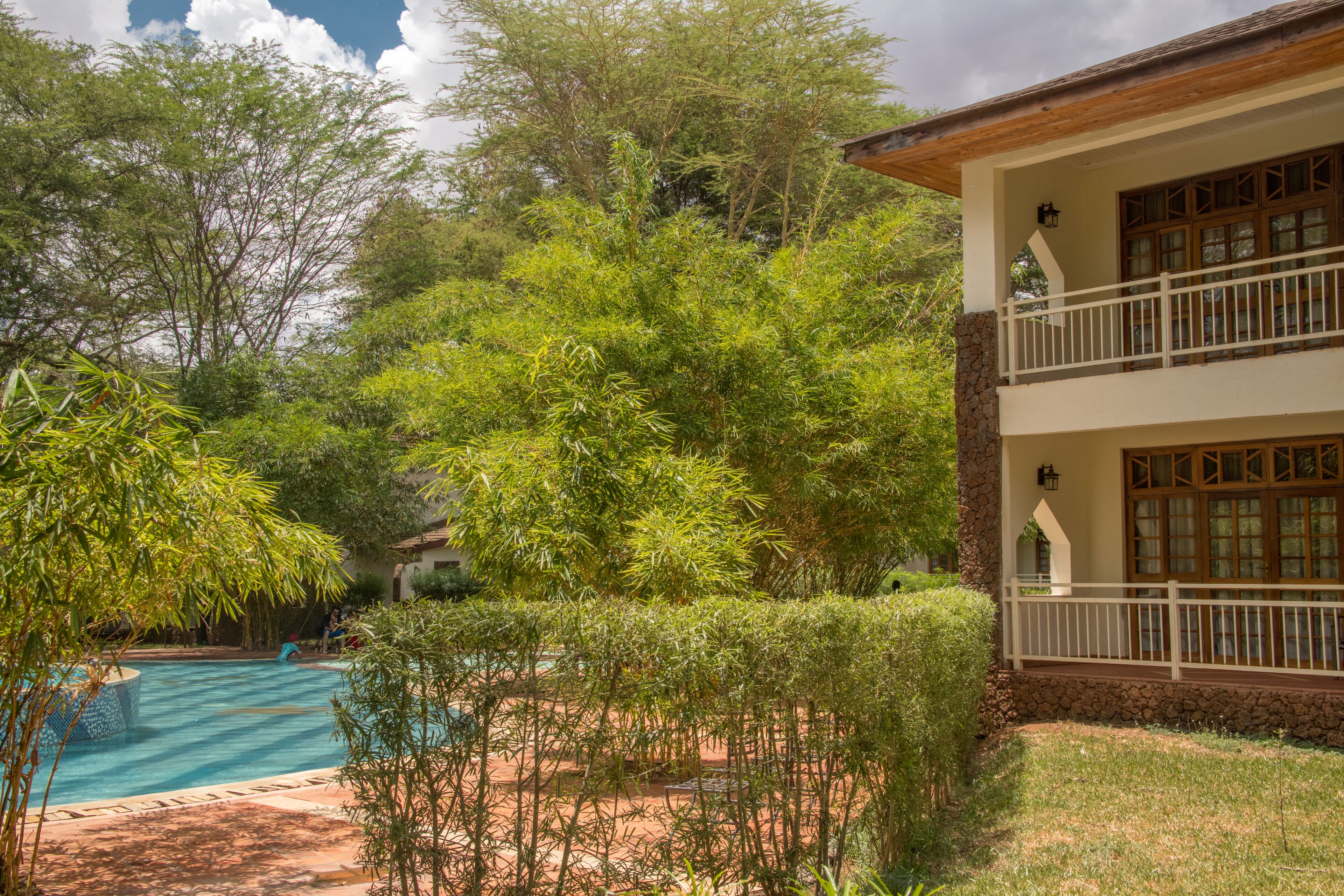 A pool surrounded by lush greenery, a two-story building with a balcony, and a stone wall.