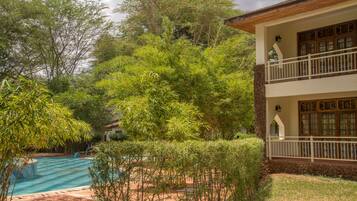 A pool surrounded by lush greenery, a two-story building with a balcony, and a stone wall.