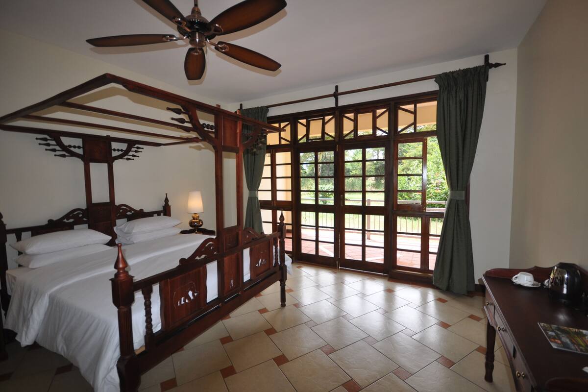 A room with a four-poster bed, a ceiling fan, large windows with curtains, and a wooden desk with a kettle.