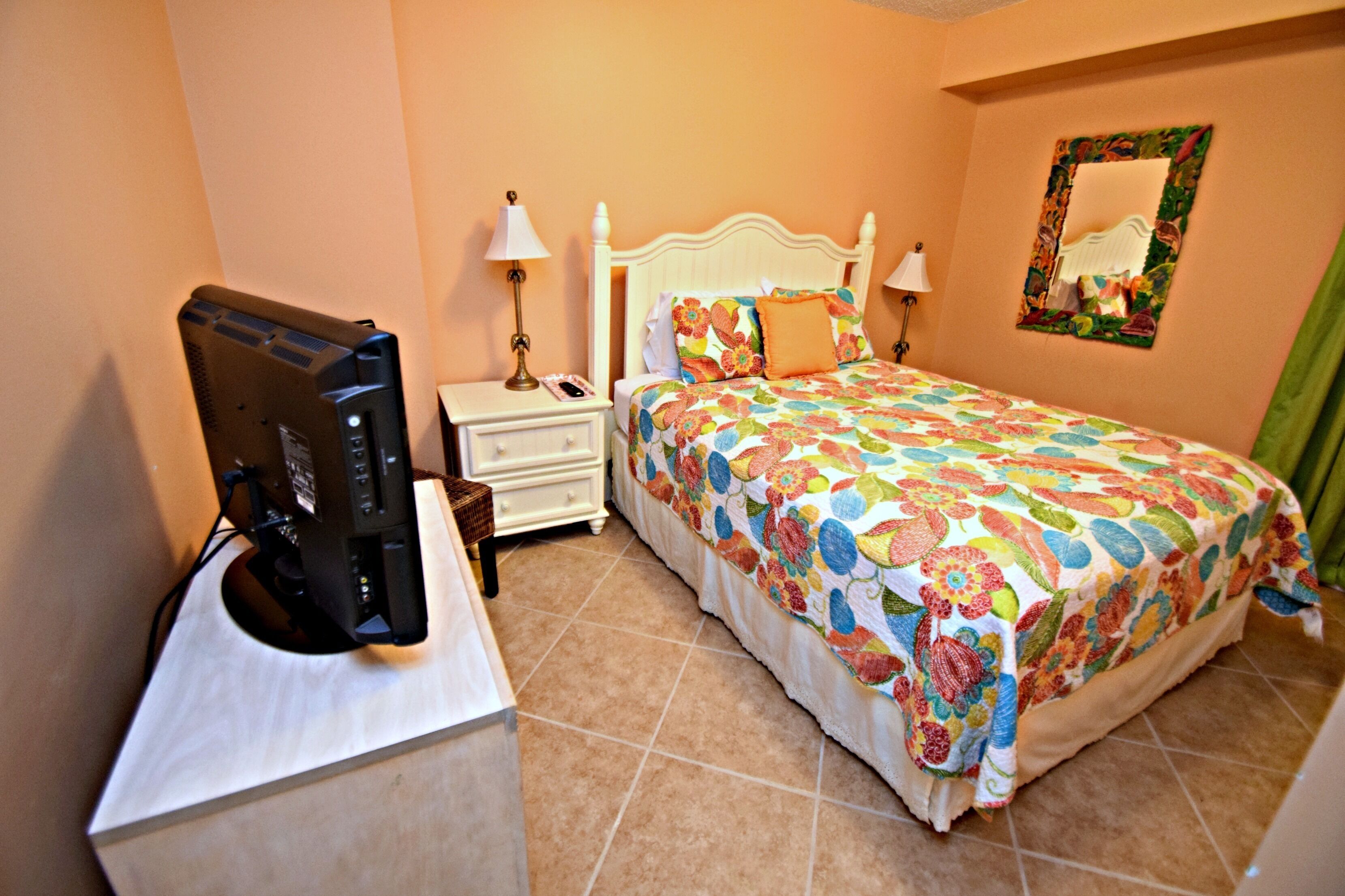 A bedroom with a bed, a nightstand, a desk with a computer, and a mirror on the wall.