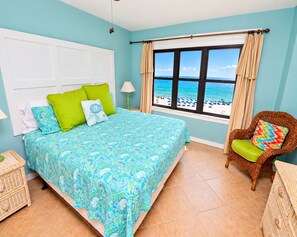 A bedroom with a bed, a chair, a window with a view of the beach, and a white door.