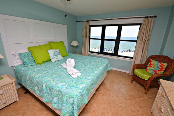 Quarto com cama, cadeira, janela vista para a praia e mesa de cabeceira.