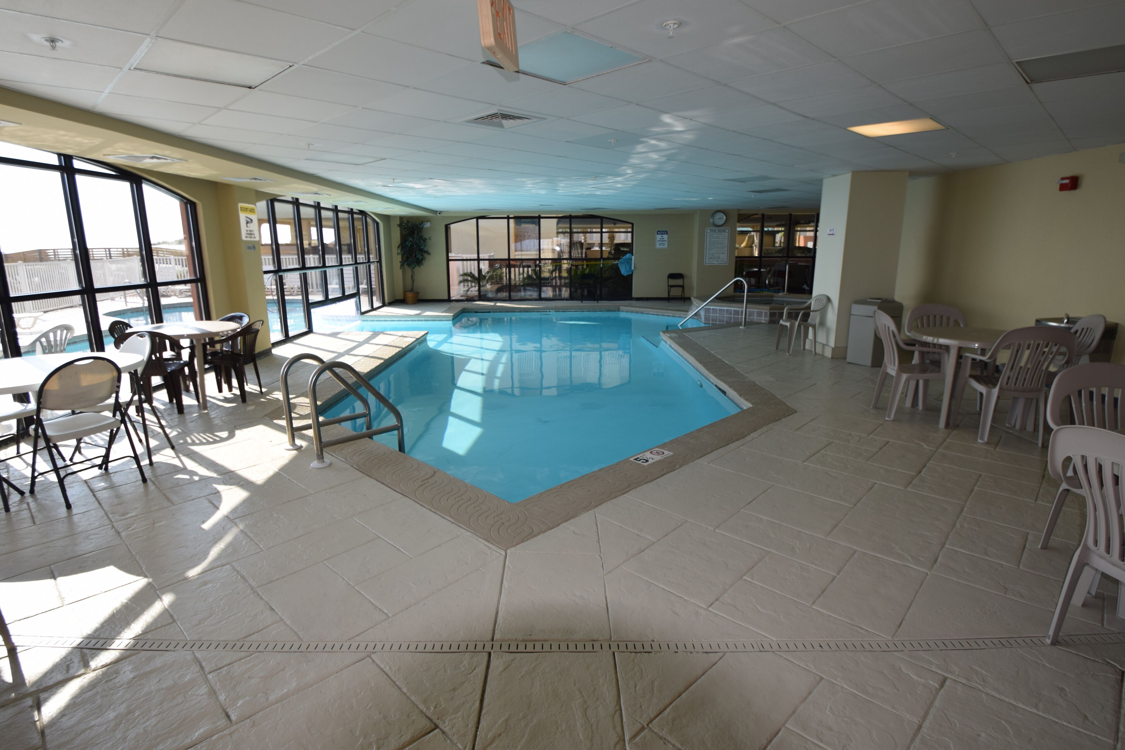 An indoor swimming pool with clear glass walls, surrounded by plastic chairs and tables.