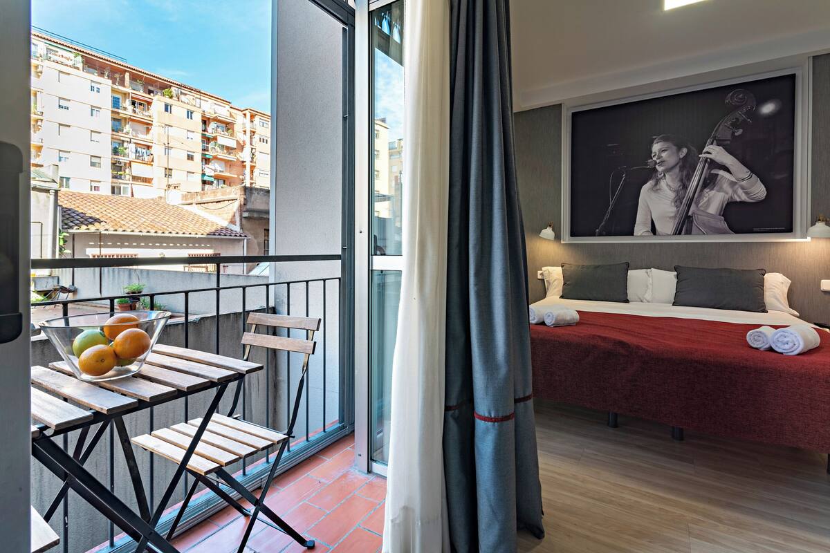 Ein Balkon mit Tisch, Stühlen und einer Schale mit Obst. Im Zimmer befindet sich ein Bett mit roter Decke und Kissen.