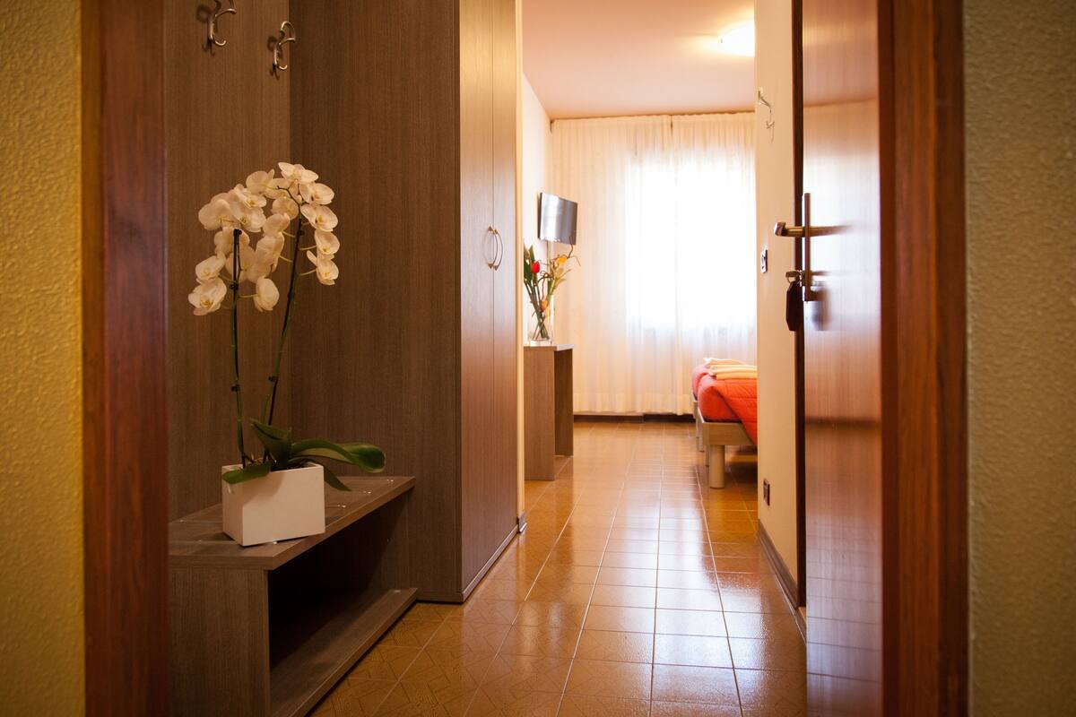 A hotel hallway with a wooden door, a potted orchid, a bedside table, and a bed with a red blanket.