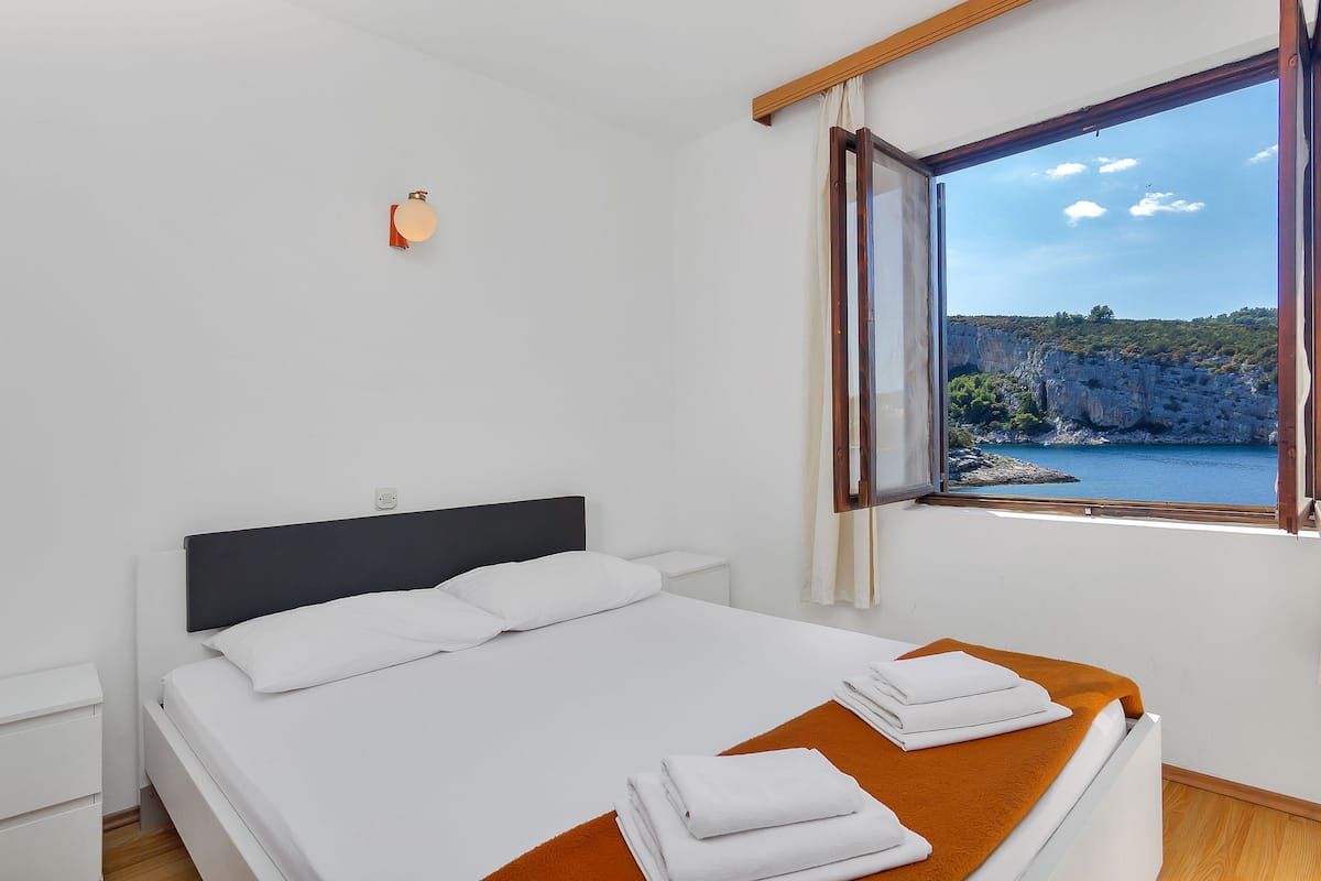 A neatly made bed with white linens and a brown throw blanket, a bedside table, and a window with a view of cliffs and water.