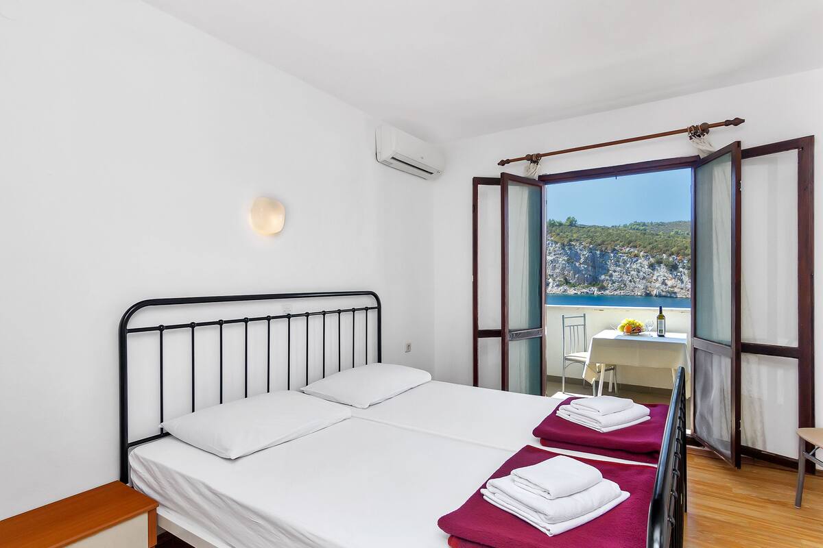 A bedroom with a bed, a nightstand, a table with a chair, and a view of a balcony with a table and chairs.