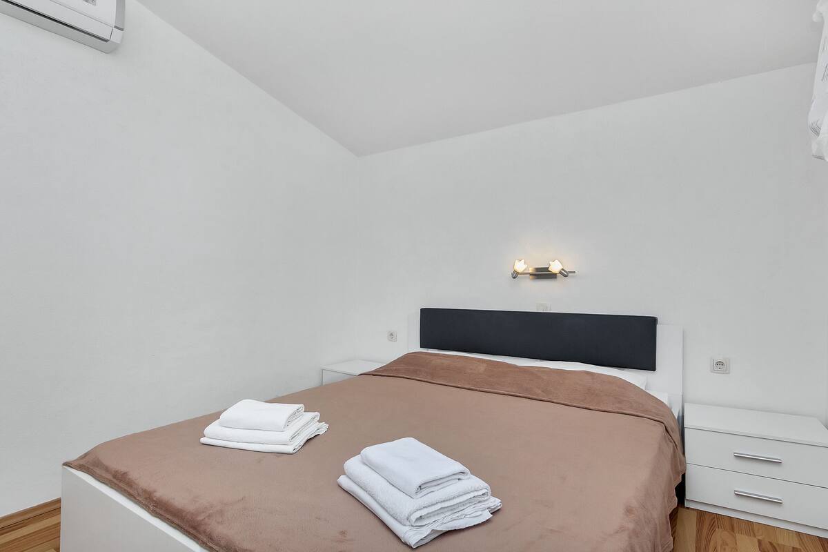 A neatly made bed with a brown bedspread and white towels, a bedside table with a lamp, and a white nightstand.