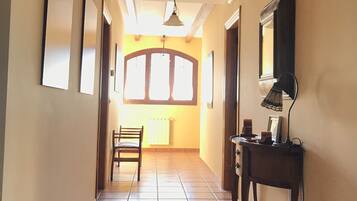 A hallway with tiled flooring, a wooden chair, a desk with a lamp, and a window with a view of the outside.