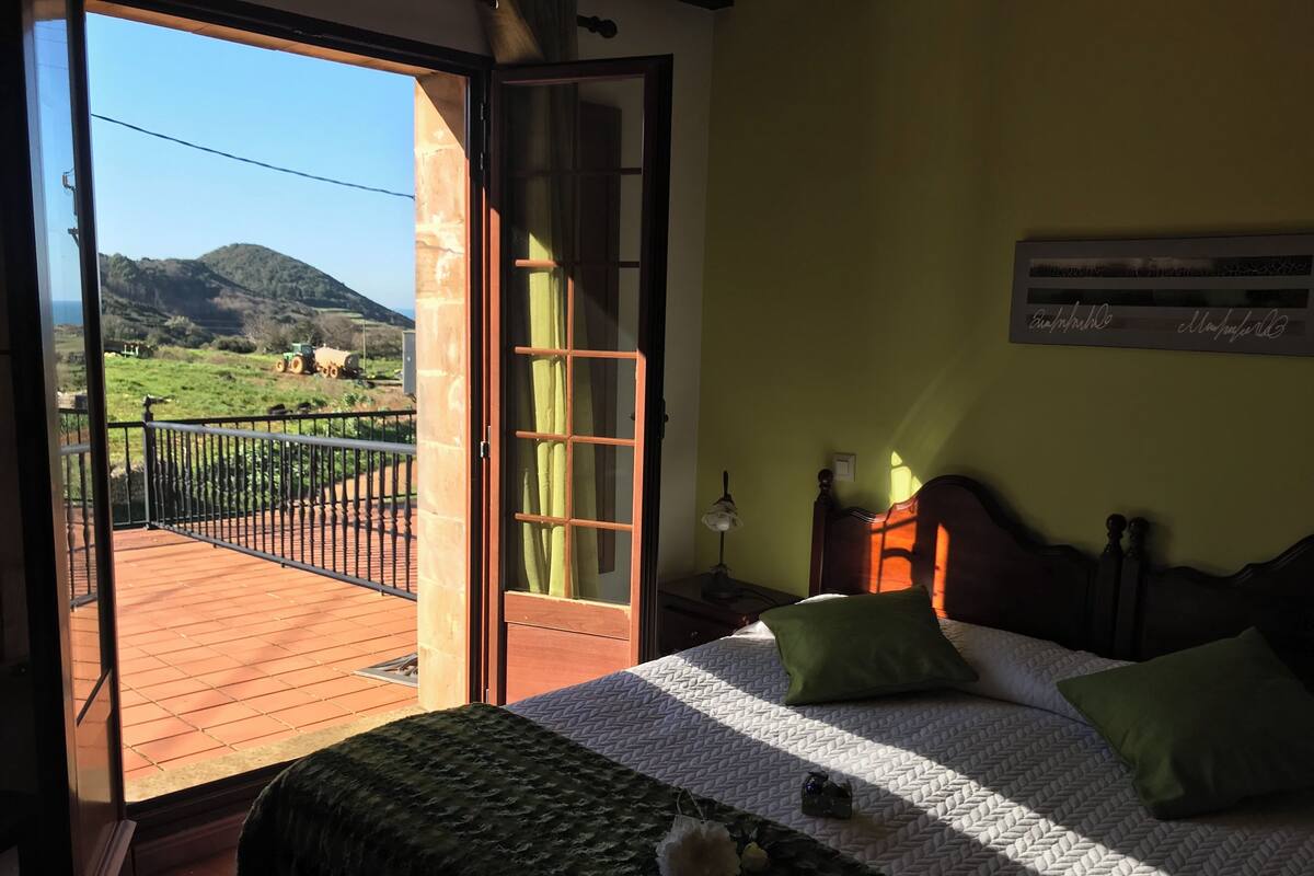 Ein Schlafzimmer mit Bett, Kissen, einem Nachttisch und einem Fenster mit Blick auf einen Balkon und Hügel.
