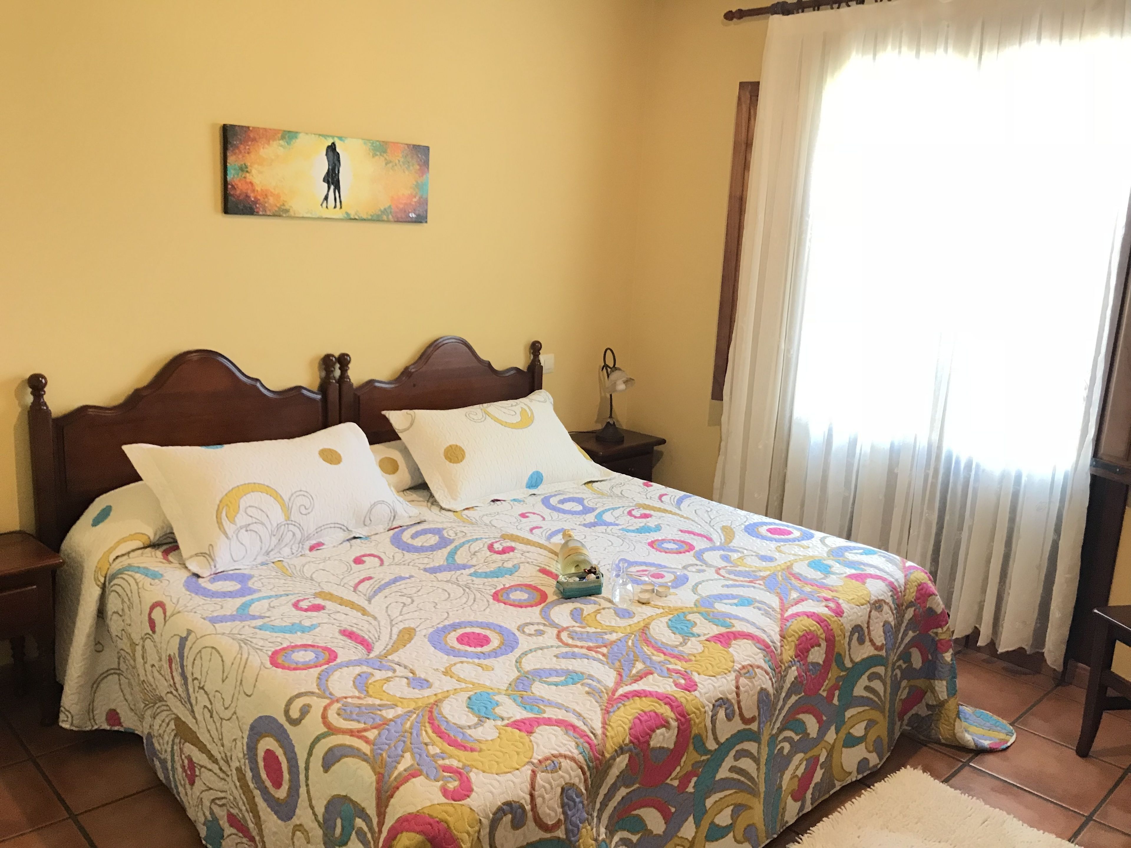 A bedroom with a wooden bed, colorful bedding, a nightstand with a lamp, a painting on the wall, and a window with curtains.